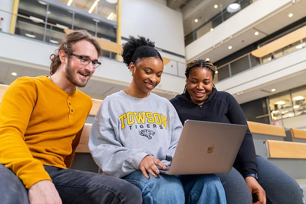 Three students are sitting together and smiling while looking at a laptop.