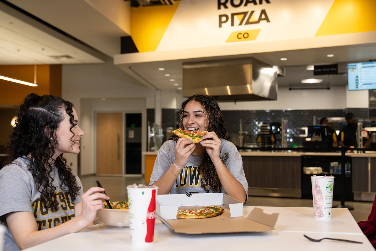 This image shows two women sitting outside of roar pizza co enjoying a slice of pizza and conversing.