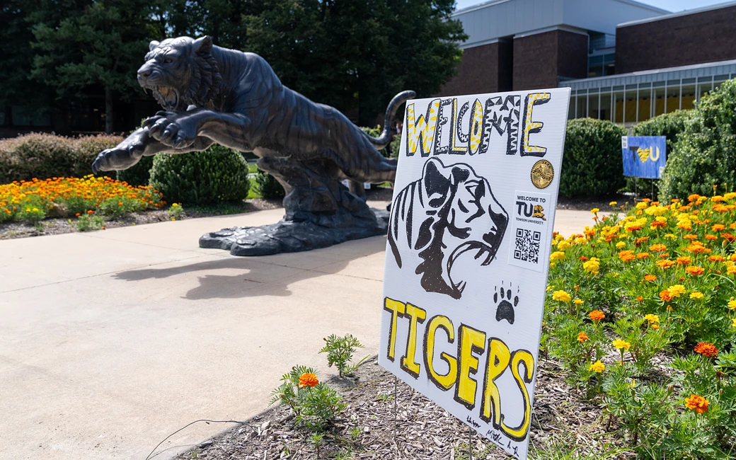 A sign reading "Welcome Tigers" is displayed in front of a tiger statue on a landscaped university campus.