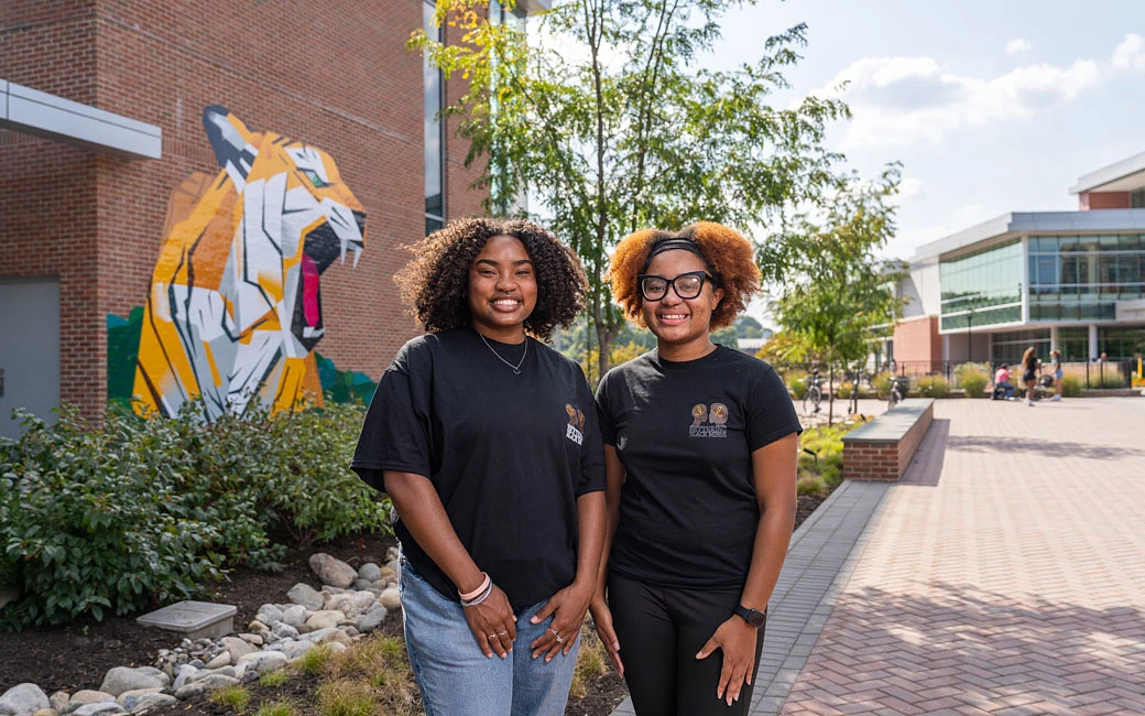 Two women standing together outdoors in front of a colorful tiger mural on a sunny day.