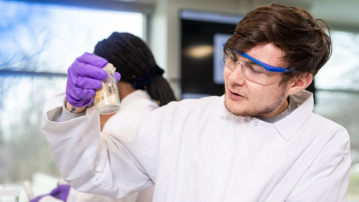 A scientist in a lab coat and safety goggles examines a sample in a bottle while wearing purple gloves.