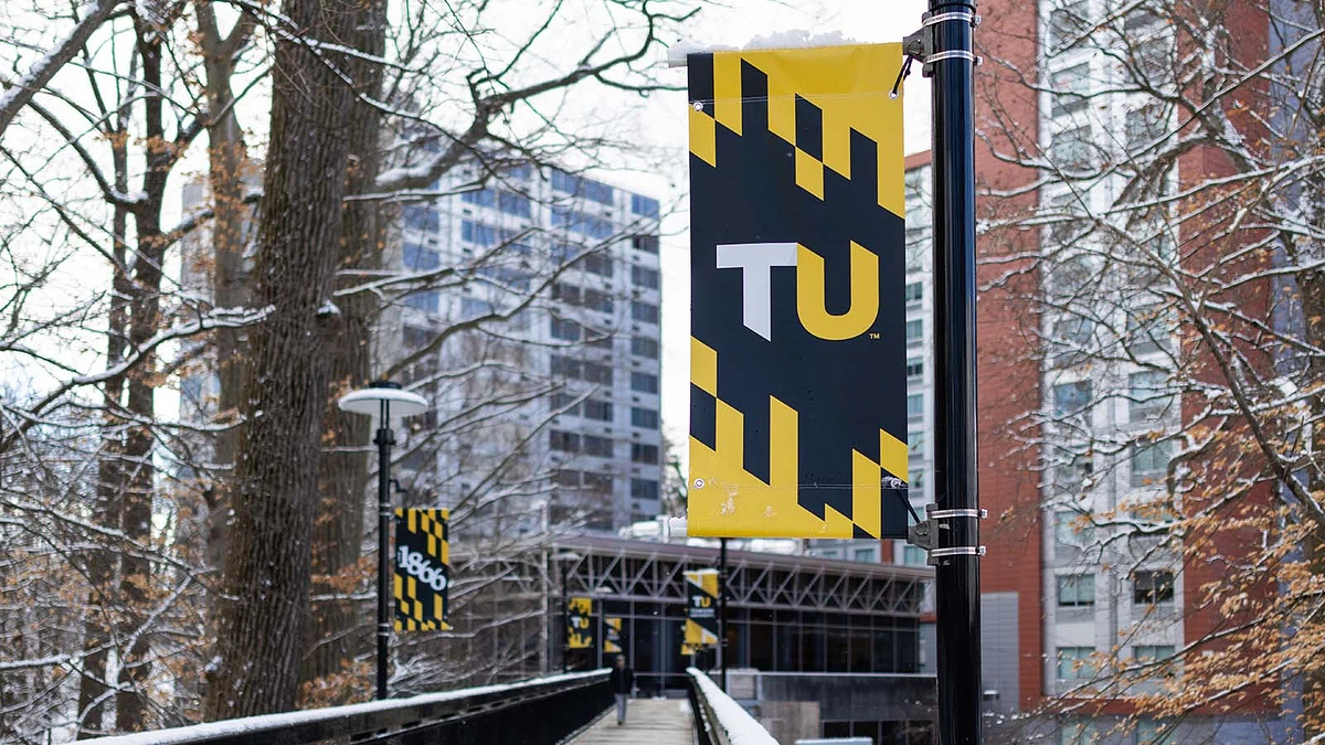 A snow-dusted campus walkway is lined with black and yellow banners displaying "TU" and "1866," with modern buildings in the background.