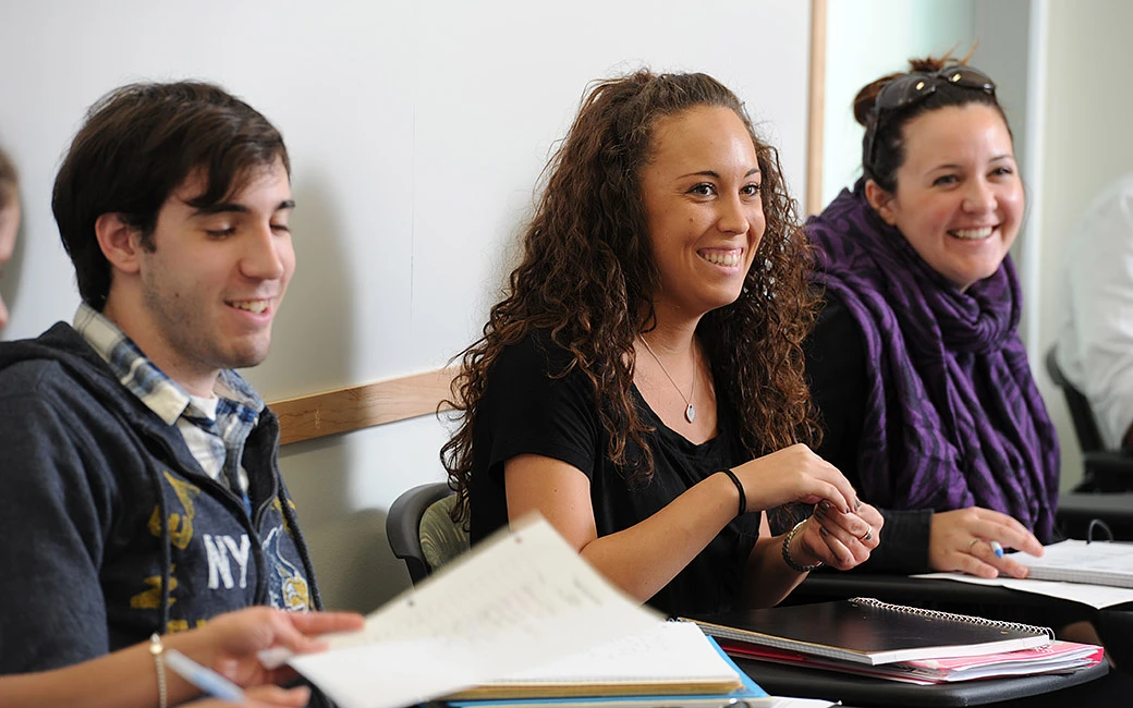 Three students are sitting in a classroom, smiling and engaging with each other while taking notes.